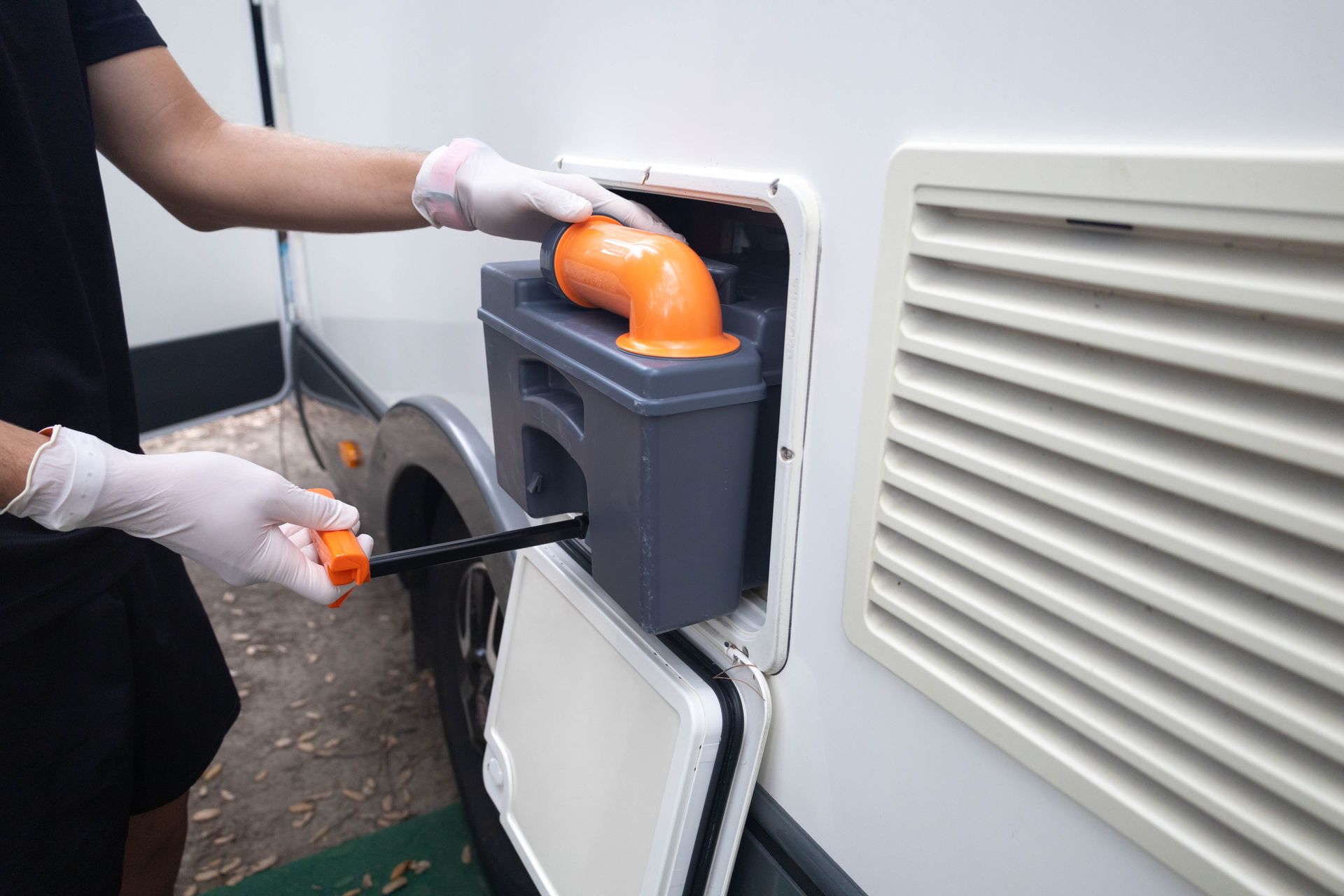 Person wearing gloves emptying a portable RV toilet cassette; white RV exterior.