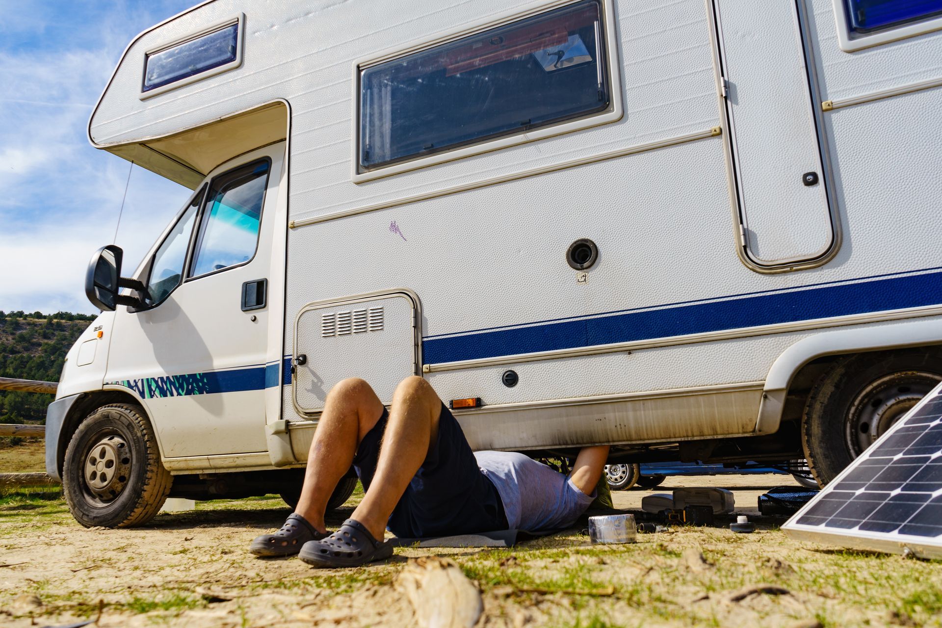 Man working on the undercarriage of a white RV, outside near a solar panel.