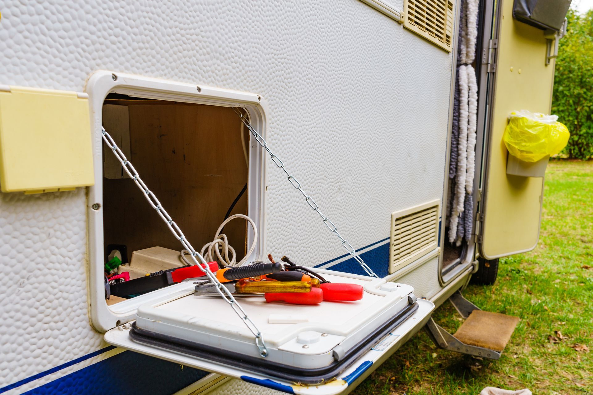 Open storage compartment on a white camper with tools visible, held by chains. Exterior shot on grass.