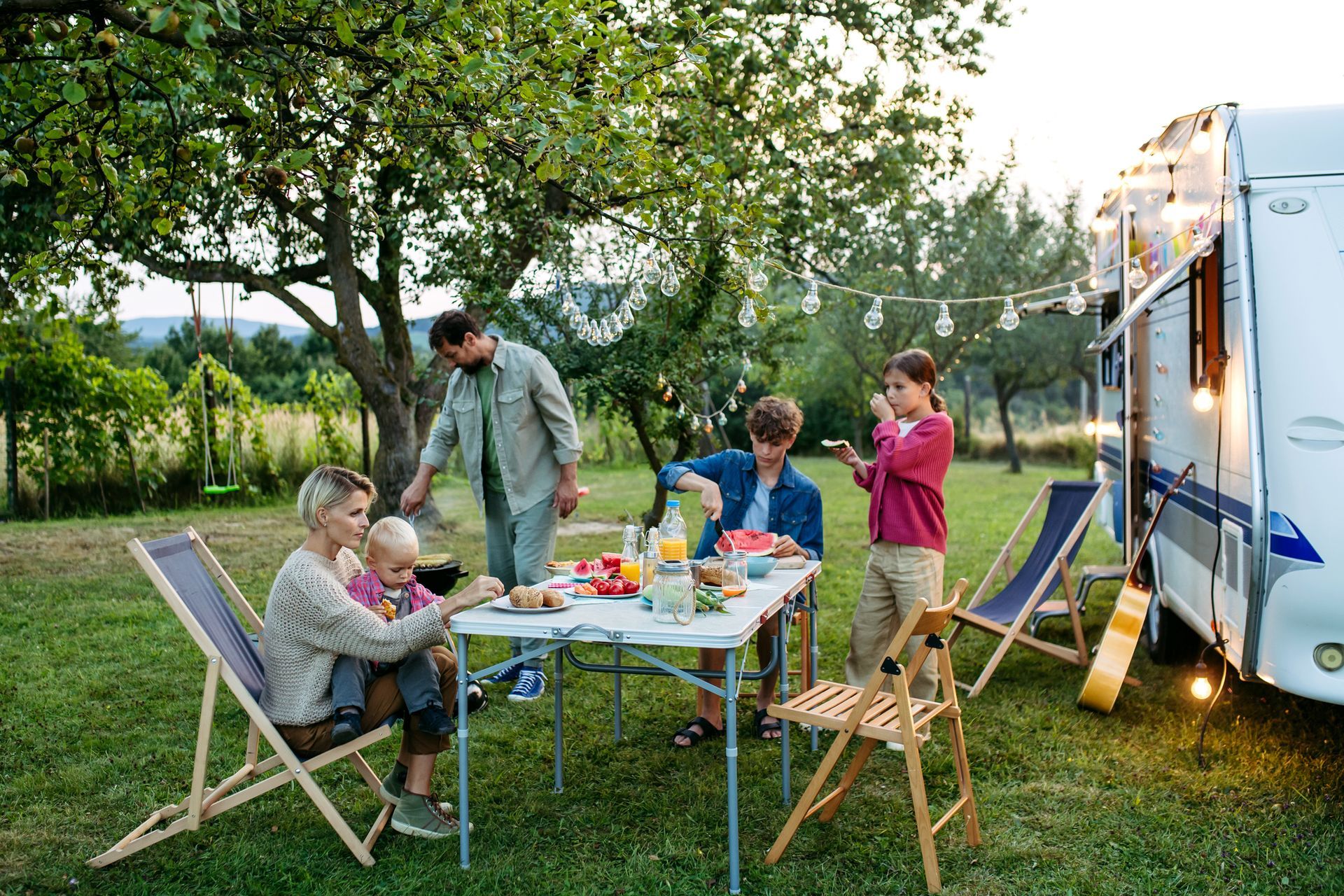 Family camping at a picnic table near an RV, eating and socializing outdoors on a sunny day. Family enjoying a picnic outdoors next to a camper, with food, drinks, and folding chairs.