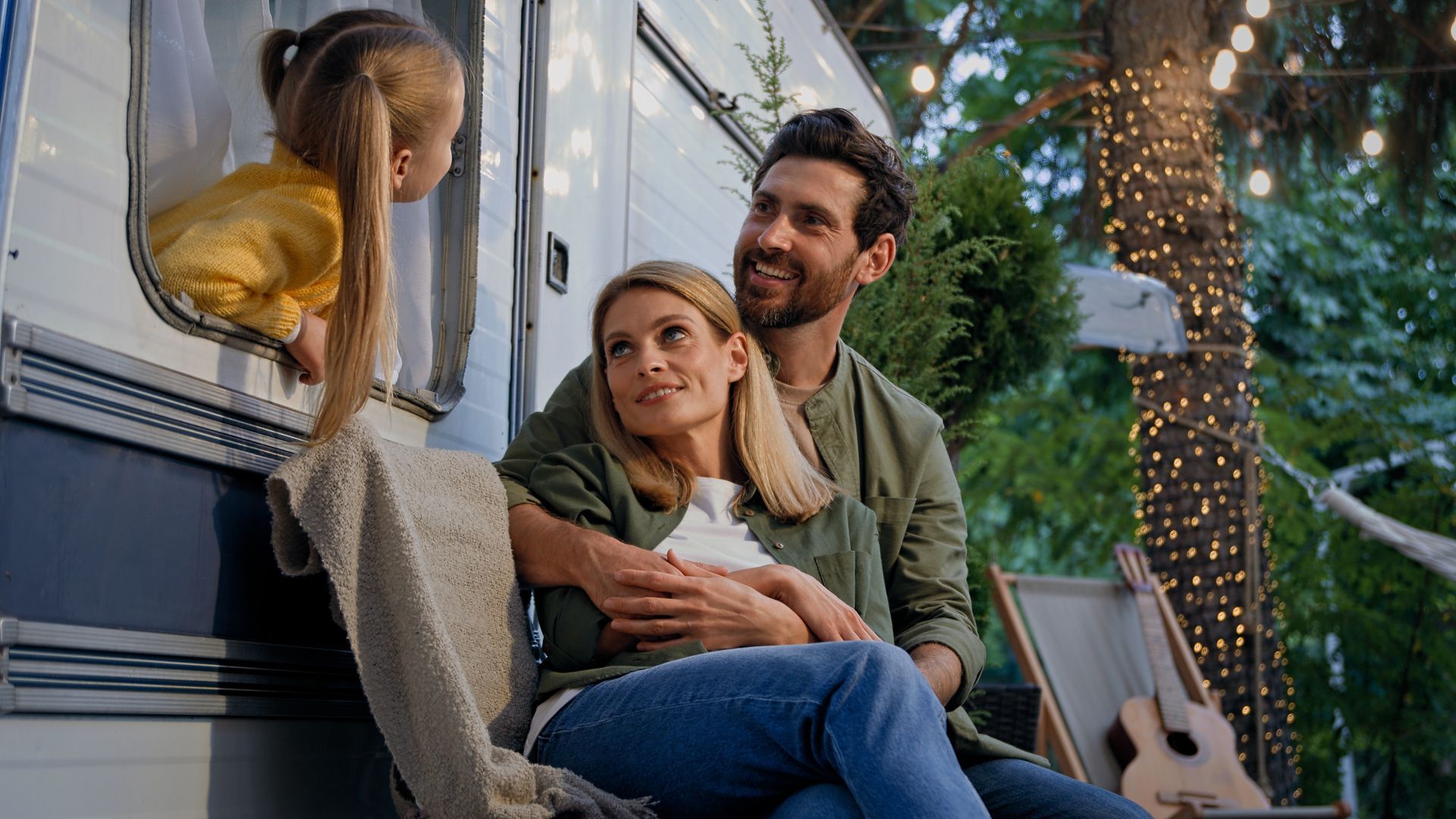Family camping by a camper: parents embrace while child looks out window, smiling. Lit with fairy lights.