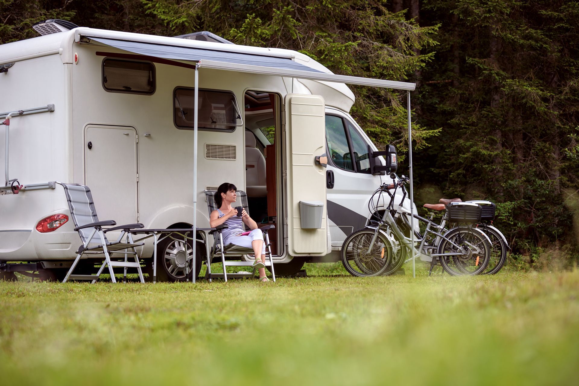 Woman relaxing outside a white RV in a grassy area, with bikes nearby.