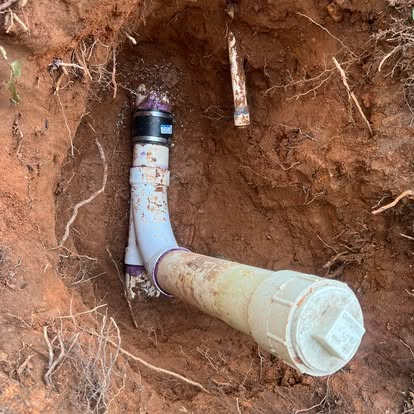 Pipes buried in dirt, connected with black and white fittings. Open hole, dirt and roots visible.