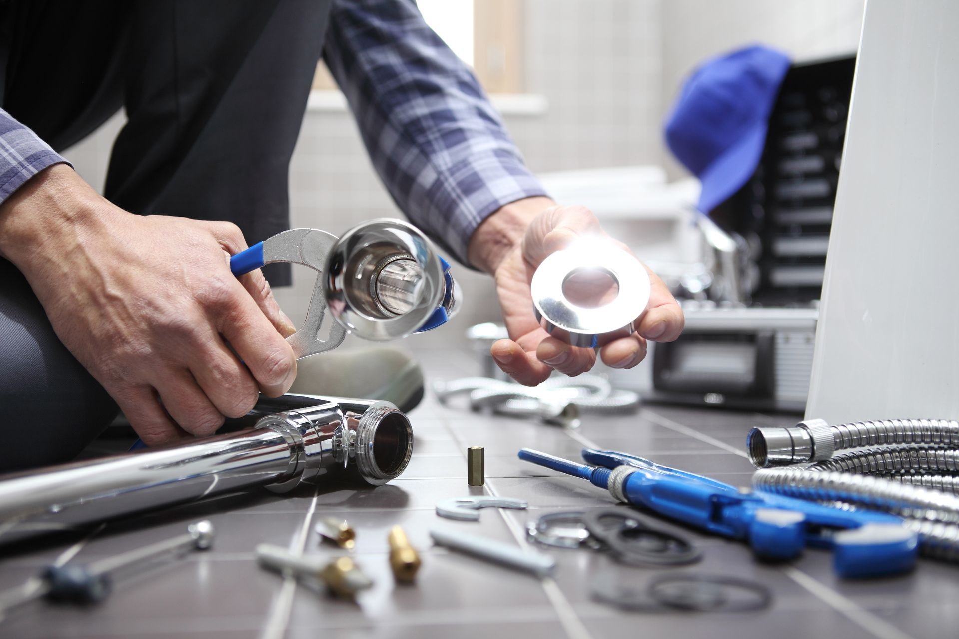 A plumber holding a metal pipe fitting and a circular escutcheon plate, with tools and plumbing components on the floor.