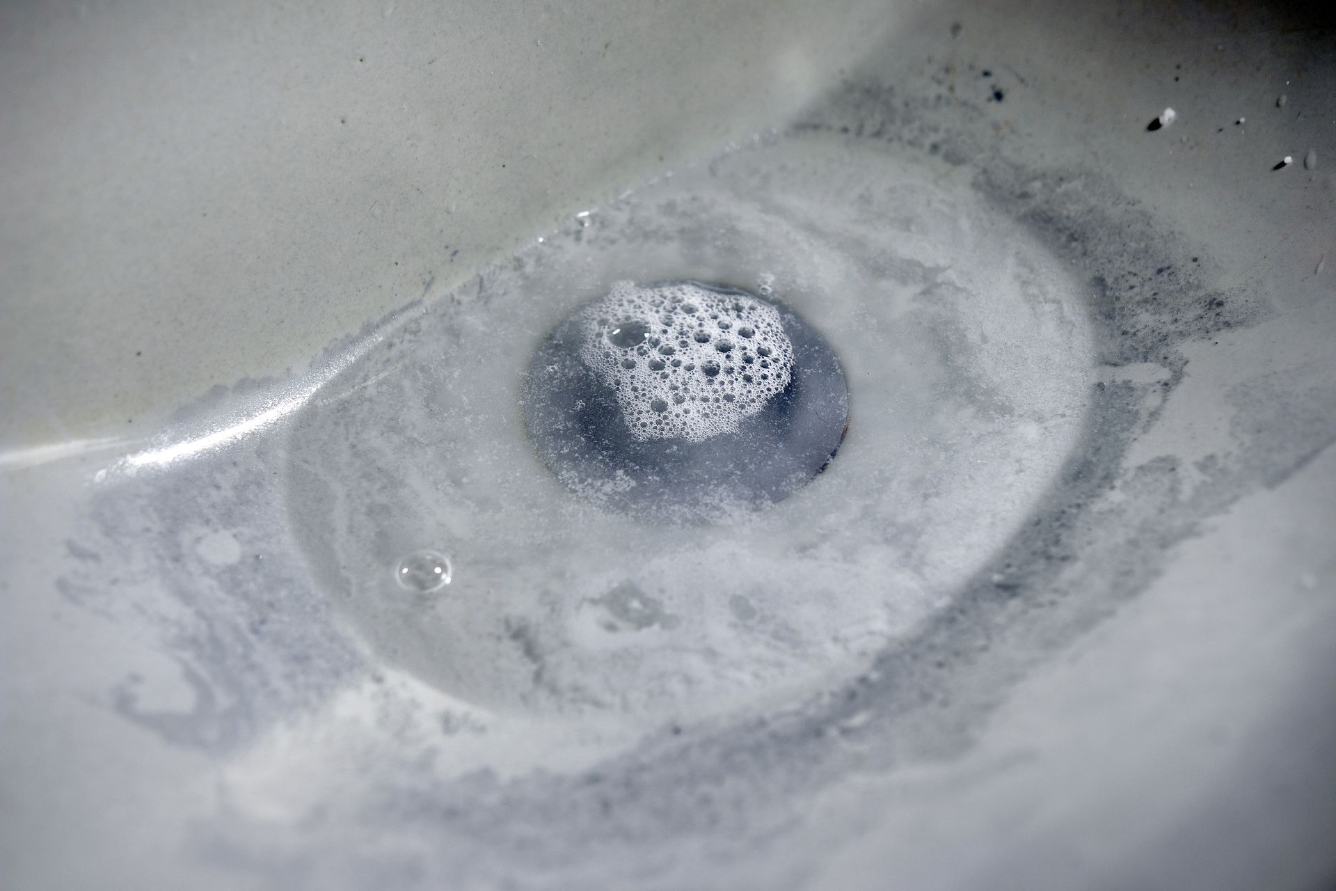 A close-up of a white bathroom sink drain with standing water, soap scum, and a small patch of bubbles.