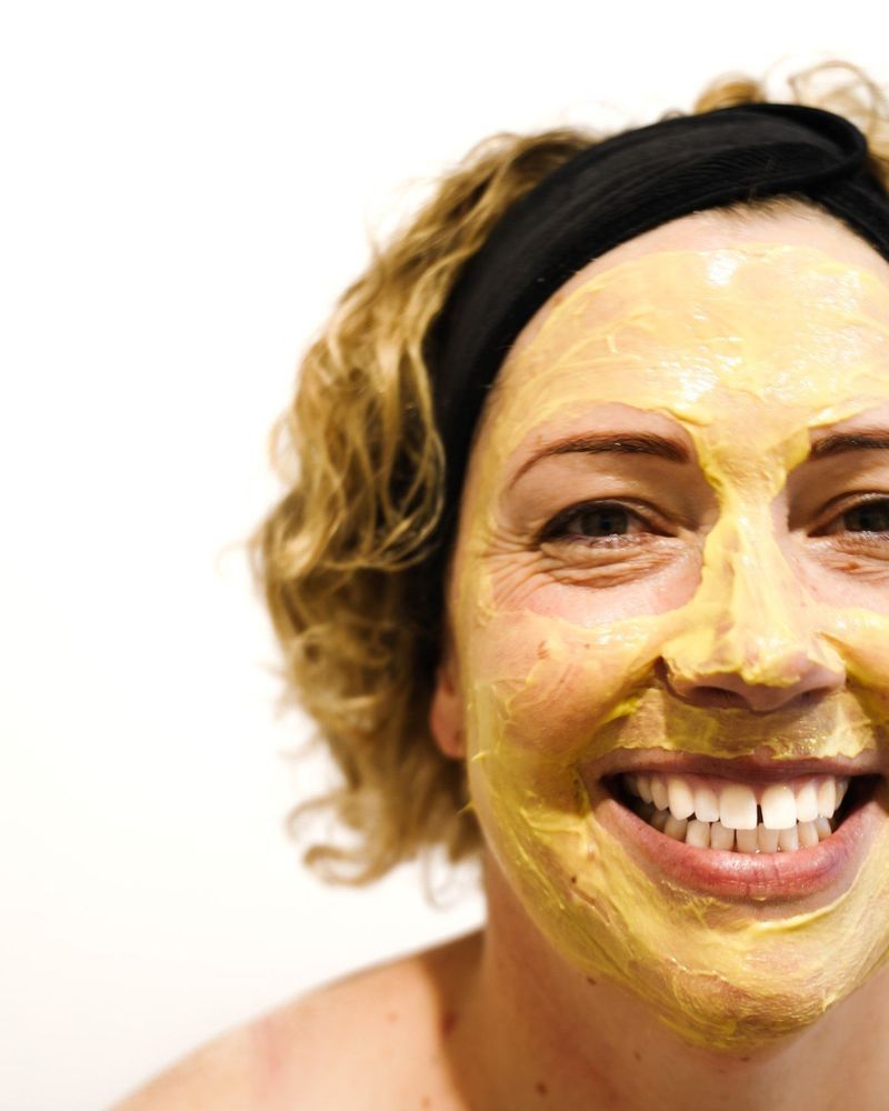 Woman smiling with yellow face mask, black headband, against white background.
