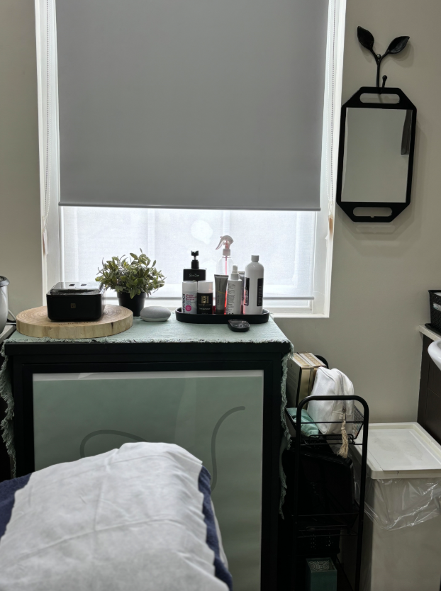 A massage table and skincare products sit by a window with a roller blind. A decorative mirror hangs on the wall.