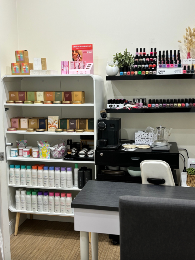Nail salon interior with shelves displaying products, a black cart with a coffee machine, and a manicure table.