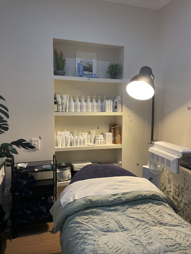 Aesthetician's treatment room with bed, products on shelves, and a lamp.