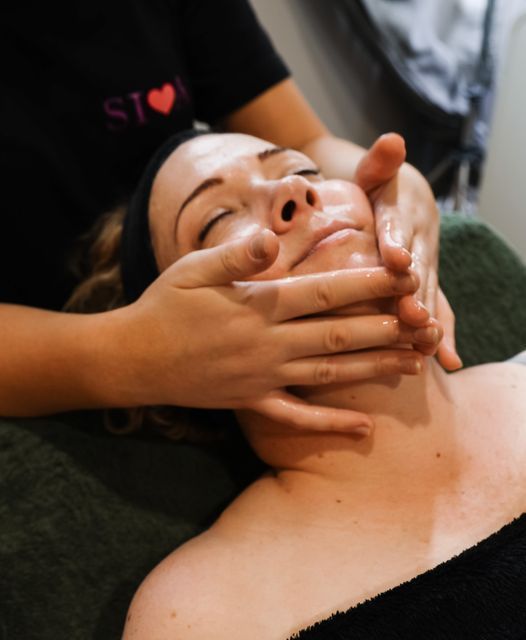 Woman receiving a facial massage with hands on her face. She has closed eyes and is lying down.