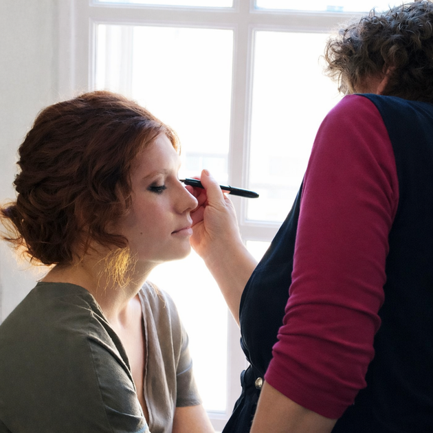 Woman getting makeup applied by a person in front of a bright window.
