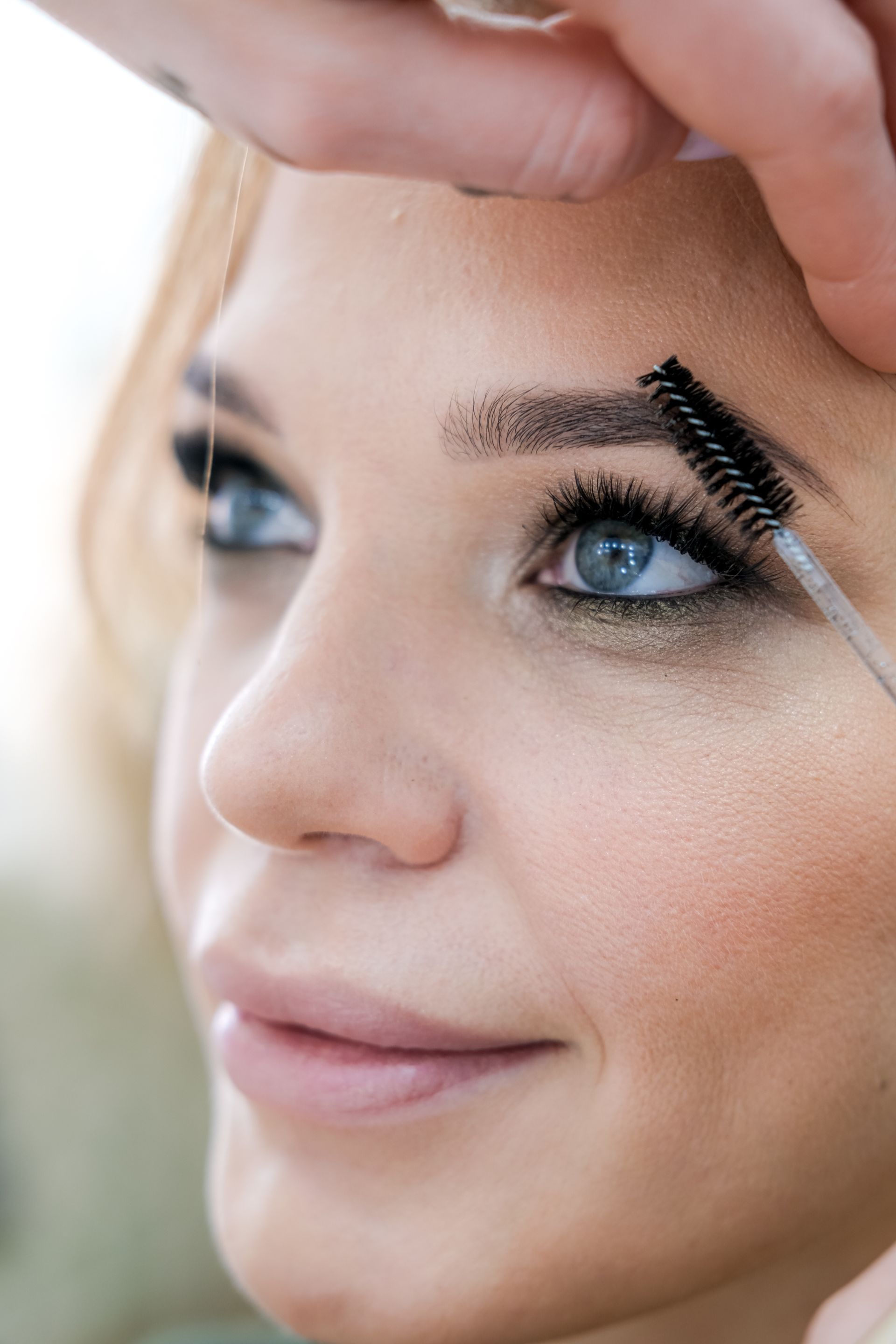 Woman's eyebrow being groomed with a brush; fair skin, light eyes, smoky eye makeup.