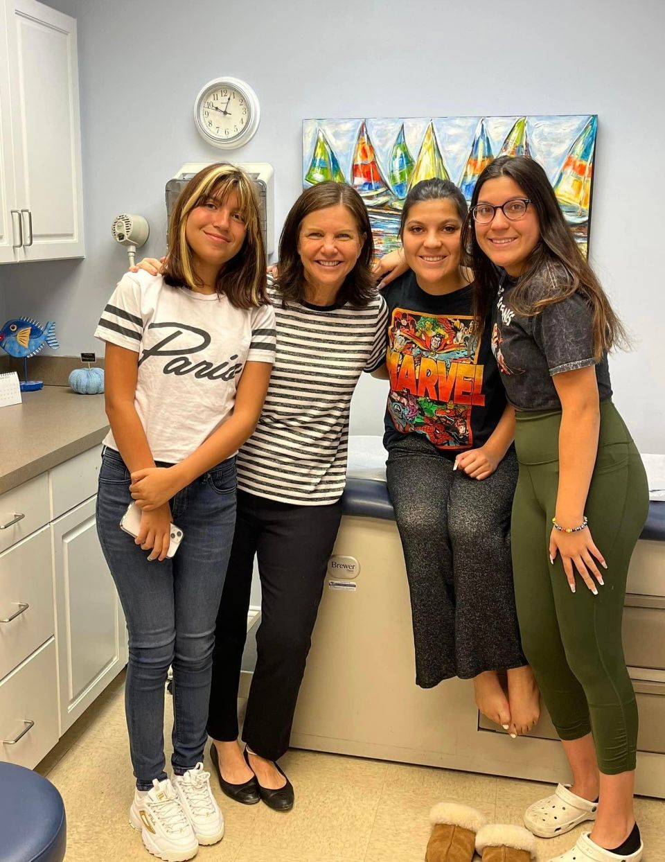 Four young women pose in a doctor's office. They are in a medical exam room in front of a table and colorful painting.