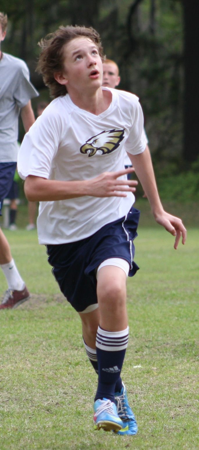 A young person in a white soccer jersey and blue shorts runs on a field, looking upward.