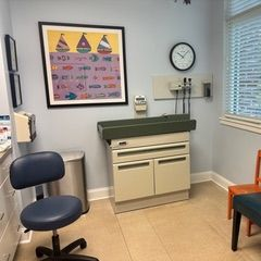 Exam room with changing table, art, clock, and medical equipment. Blue and orange chairs are present.