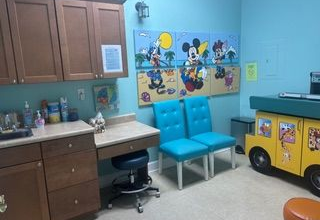 Pediatrician's office. Mickey Mouse artwork above teal chairs. Yellow school bus playset. Brown cabinets.