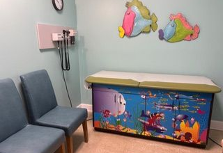 Pediatrician's exam room with blue chairs, exam table, and decorative fish on the wall.