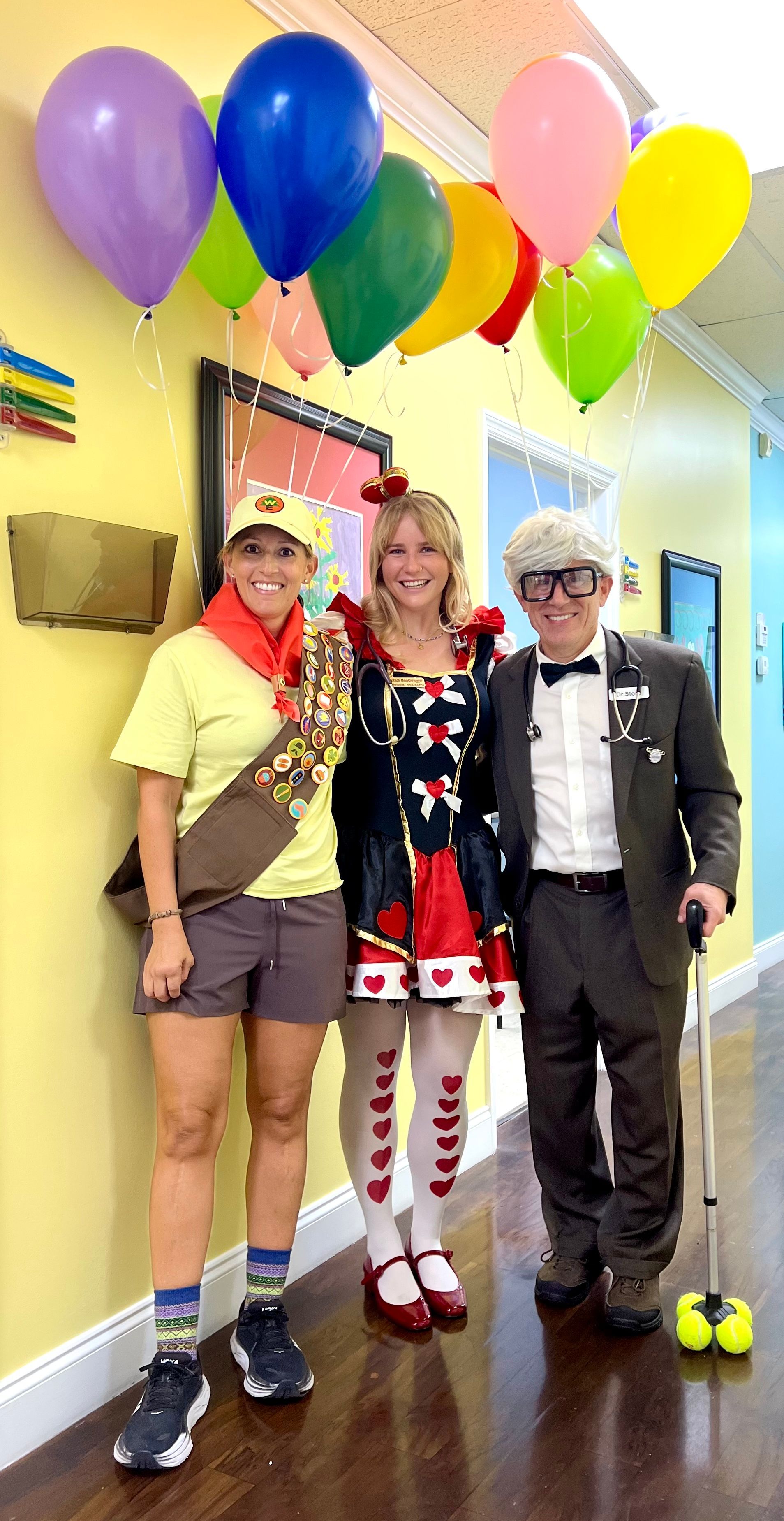 Three people in costume: Scout, Queen of Hearts, and an elderly man, under balloons.