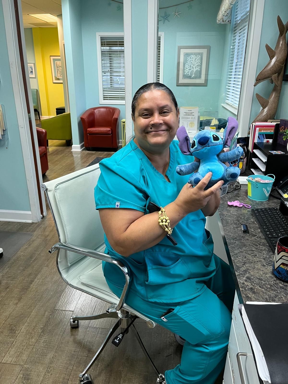 Woman in blue scrubs holding a Stitch stuffed animal, smiling, sitting at a desk in a medical office setting.