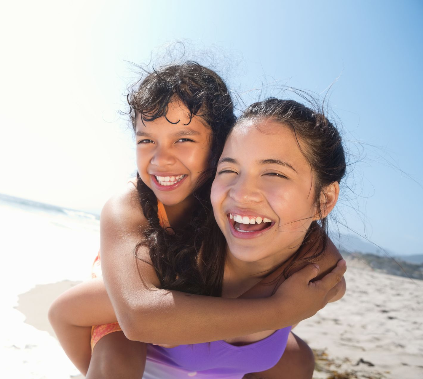 Two smiling people on a beach. One person is piggybacking the other, both looking at the camera. Sunny day.