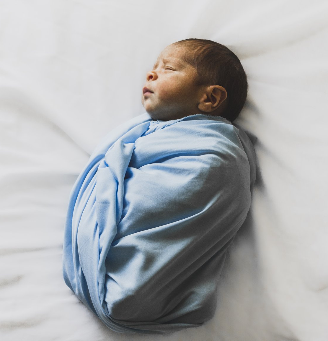 Baby swaddled in blue blanket, lying on white sheet, eyes closed.