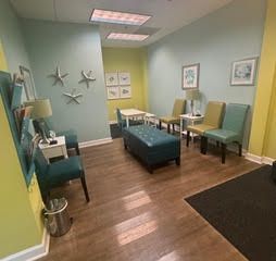 Reception area with chairs, a table, and a blue ottoman. Pale walls with artwork.
