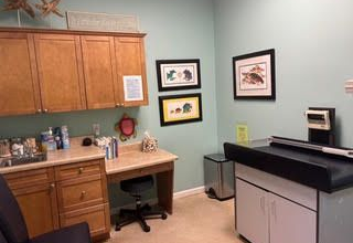 Medical exam room with wooden cabinets, desk, and examination table; light blue walls.