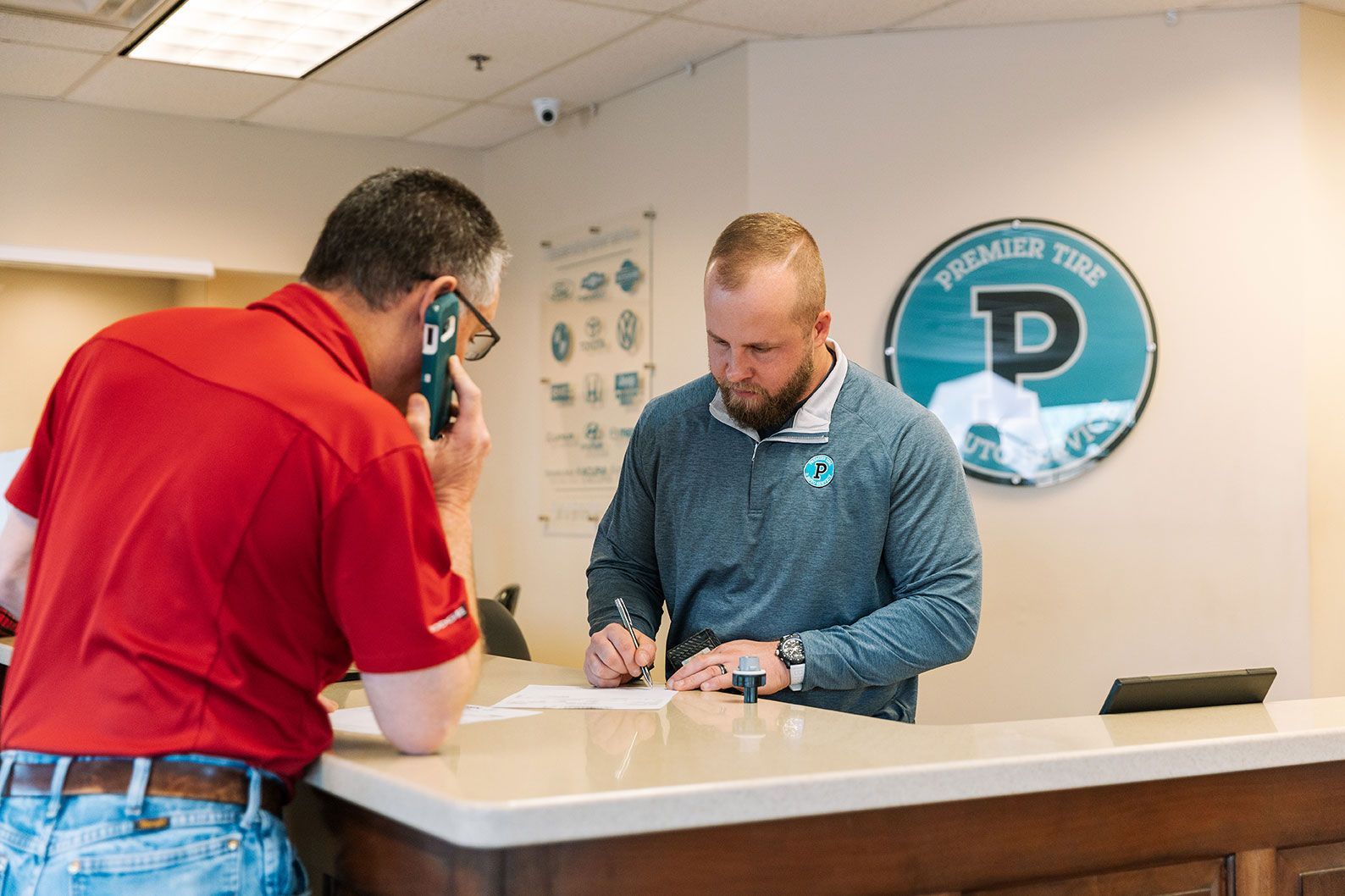 A man is signing a document while another man talks on a cell phone at Premier Tire and Auto Service