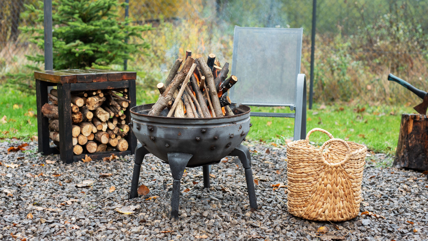 Fire pit with stacked wood and kindling outdoors; chair and basket present.
