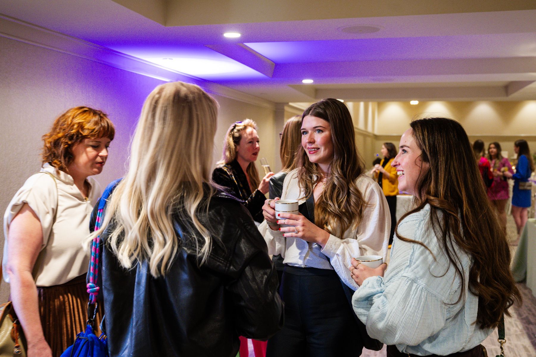 People conversing at an event. Several women are in a room with a purple lit ceiling.