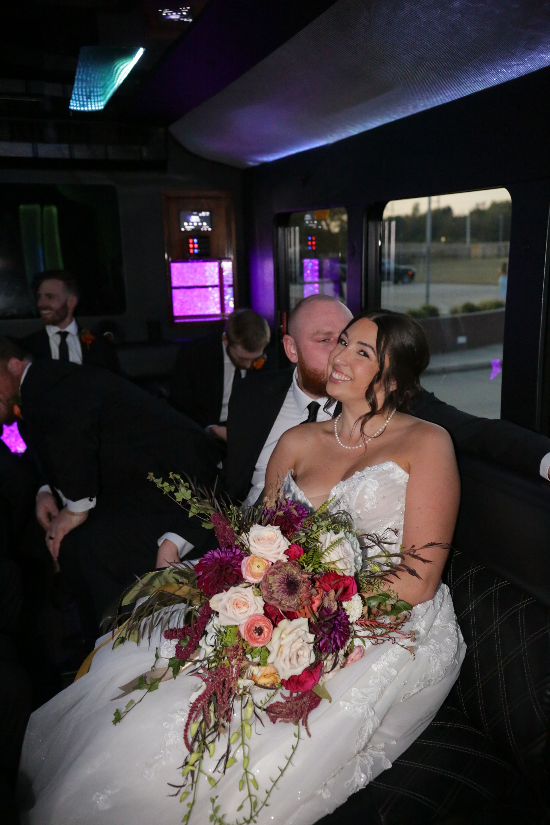 Bride and groom in a limousine, she holds a bouquet, he kisses her cheek, others in suits.