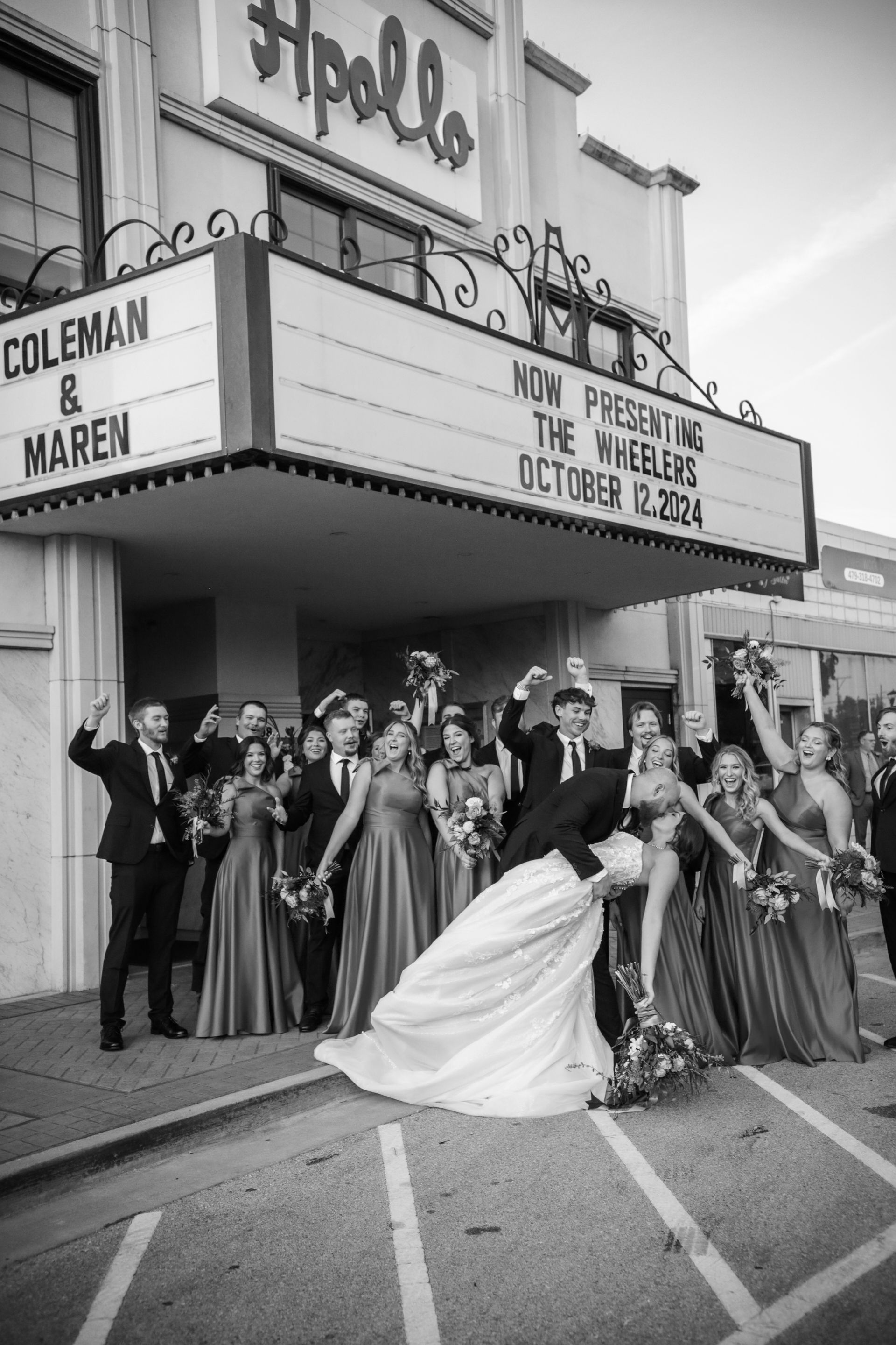 Wedding party posing in front of the Apollo Theater. Bride and groom kiss, guests cheer. Black and white.