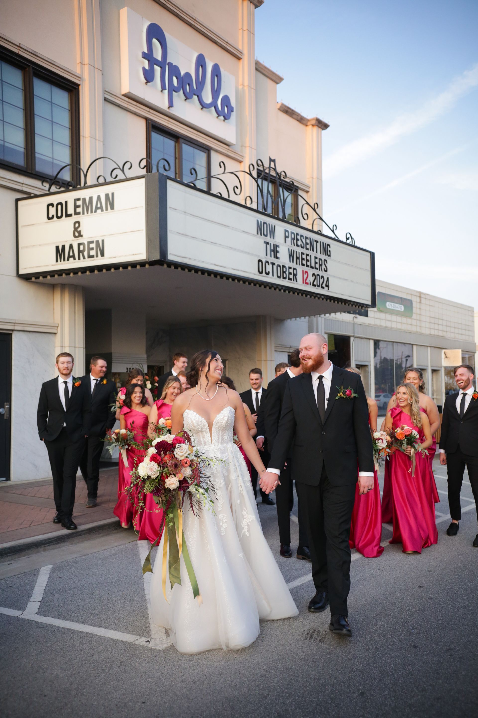 Bride and groom walking in front of a theater, holding hands. Wedding party wears dark suits and magenta dresses.