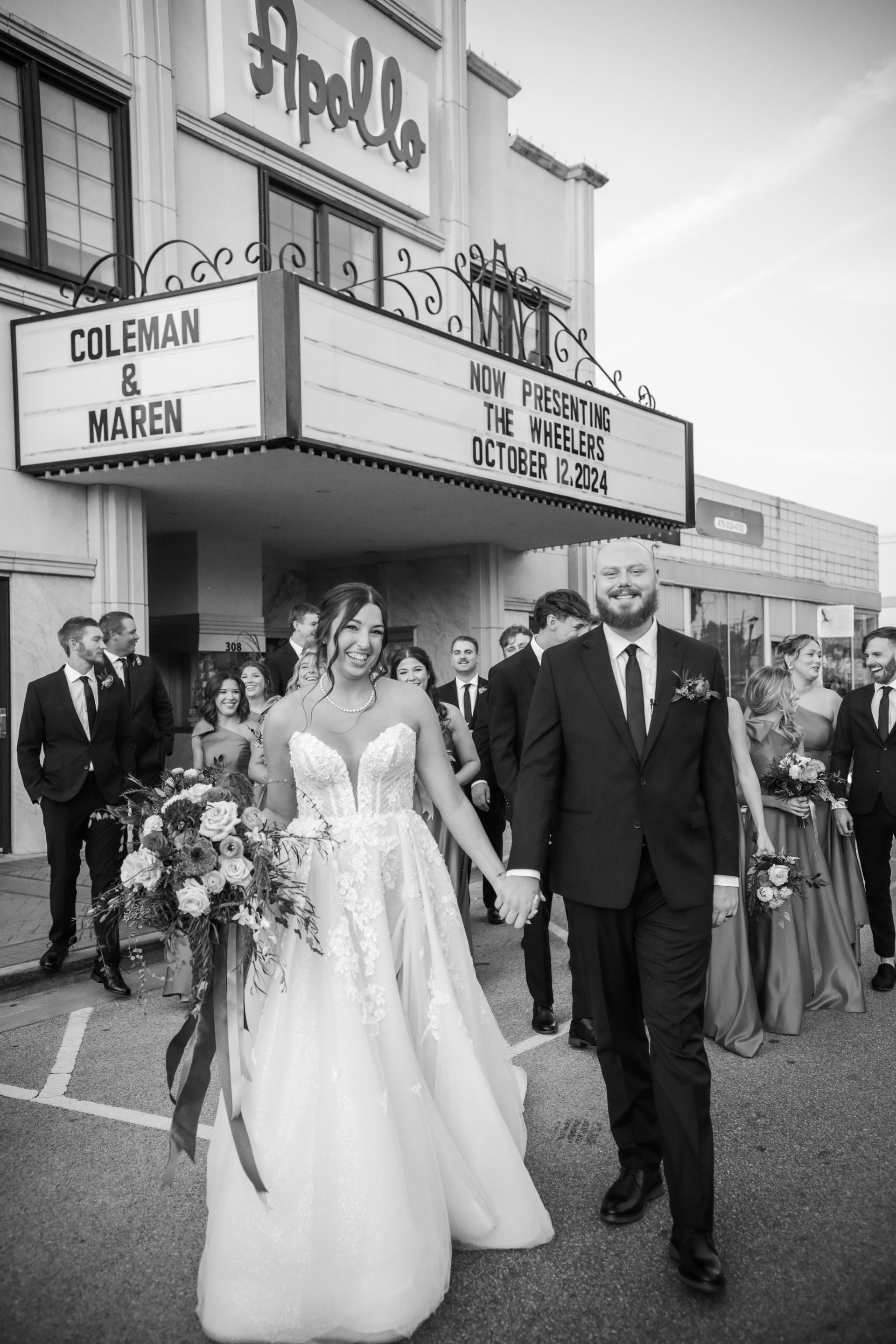 Bride and groom holding hands, exiting venue with wedding party. Apollo Theater marquee in background. Black and white.