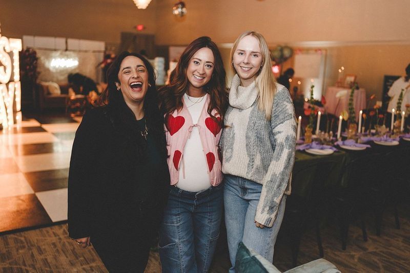 Three women smiling at a party; pregnant woman in pink sweater and jeans stands between two others.