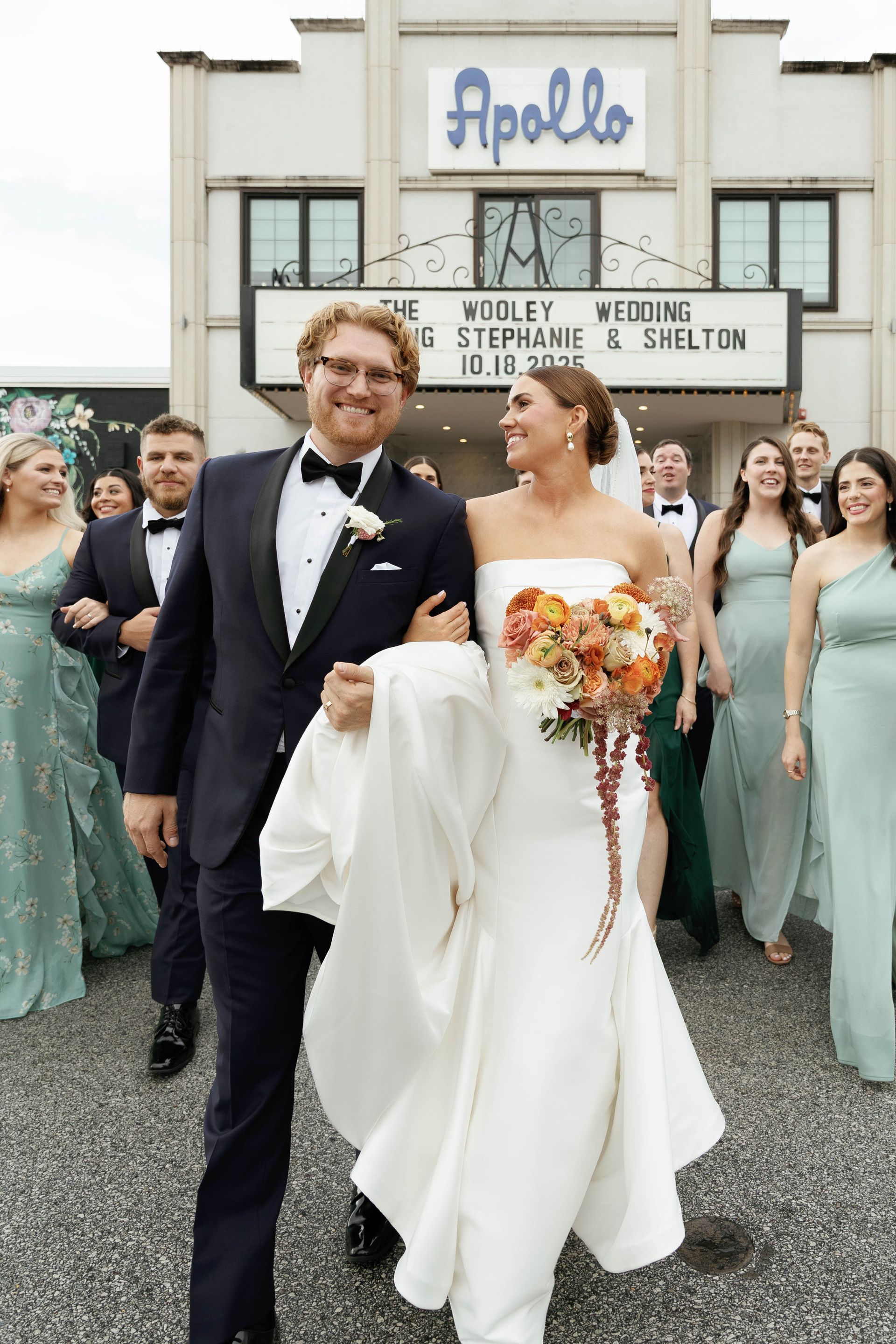 Newly married couple exits Apollo Theater with wedding party. Bride in white dress, groom in suit.