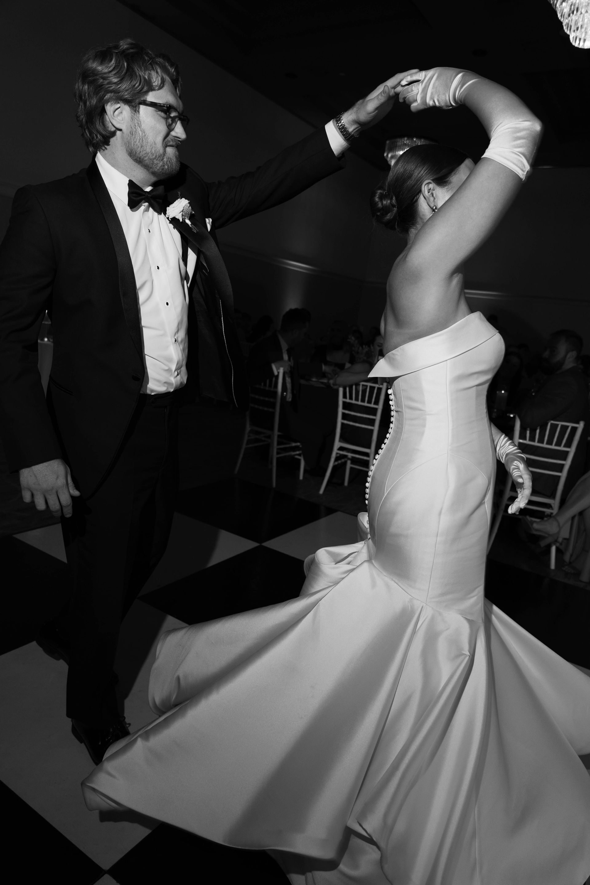 Couple dancing at a wedding reception; the bride in a mermaid gown, the groom in a tuxedo, on a checkered dance floor.