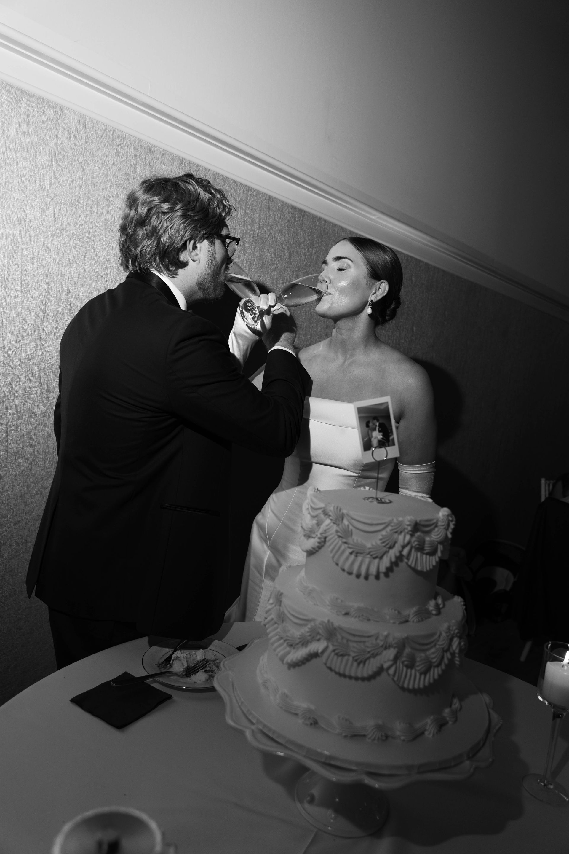 Couple toasting with champagne glasses at a wedding. Three-tiered cake on table.