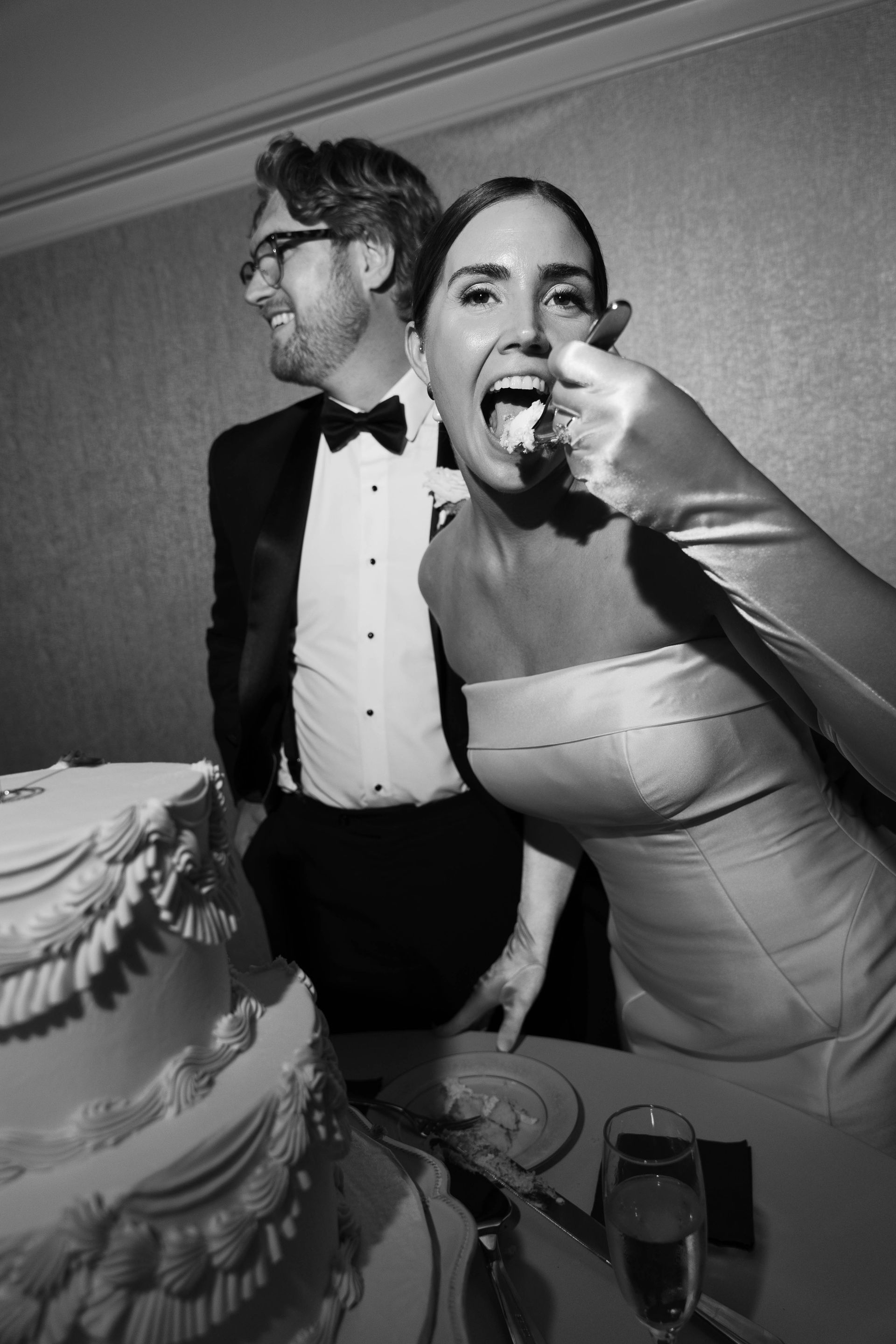 Bride eating cake, groom smiles. Wedding cake and champagne glasses on table.