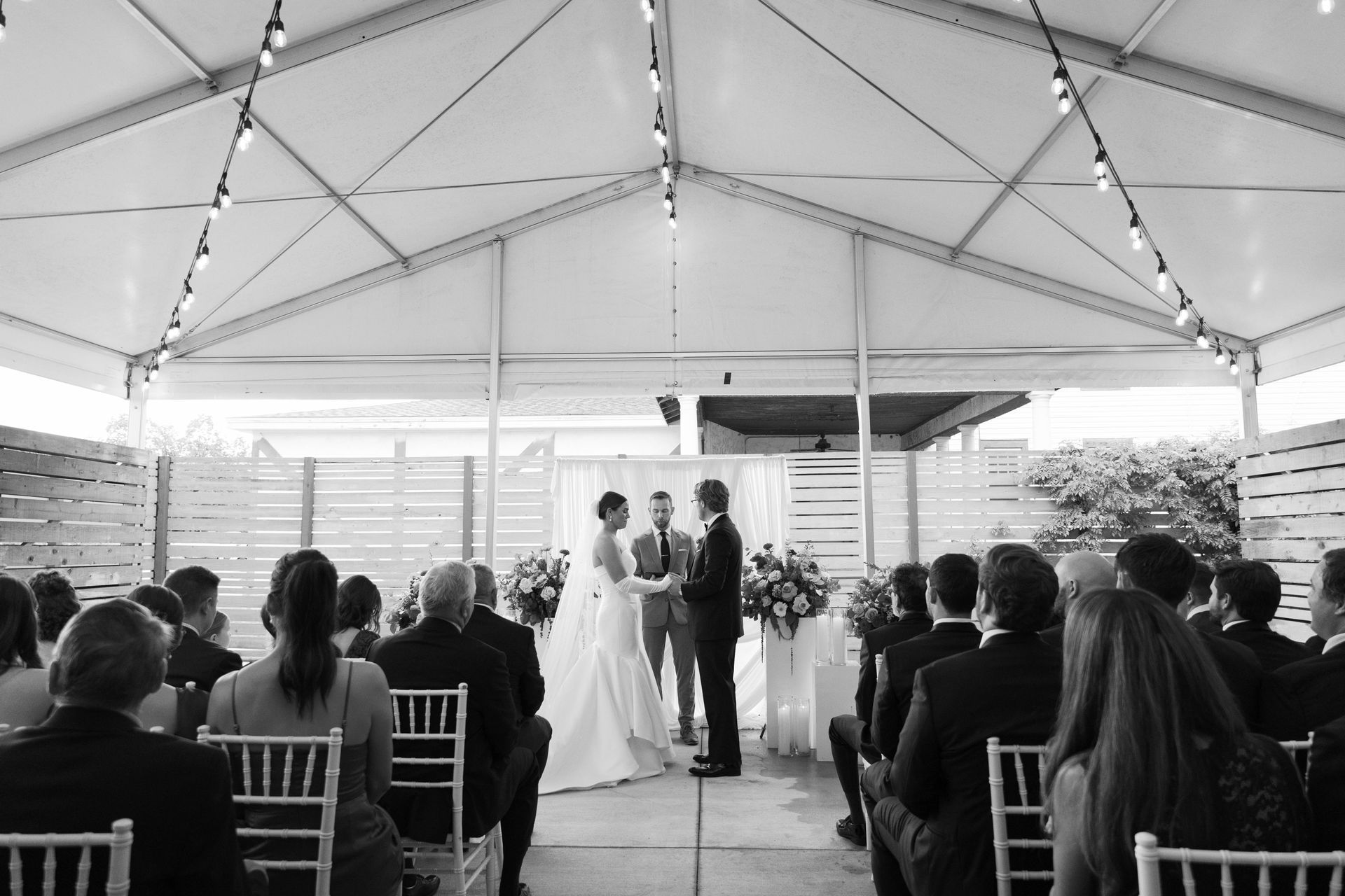 Wedding ceremony under a tent with couple exchanging vows. Guests seated in chairs, string lights overhead.