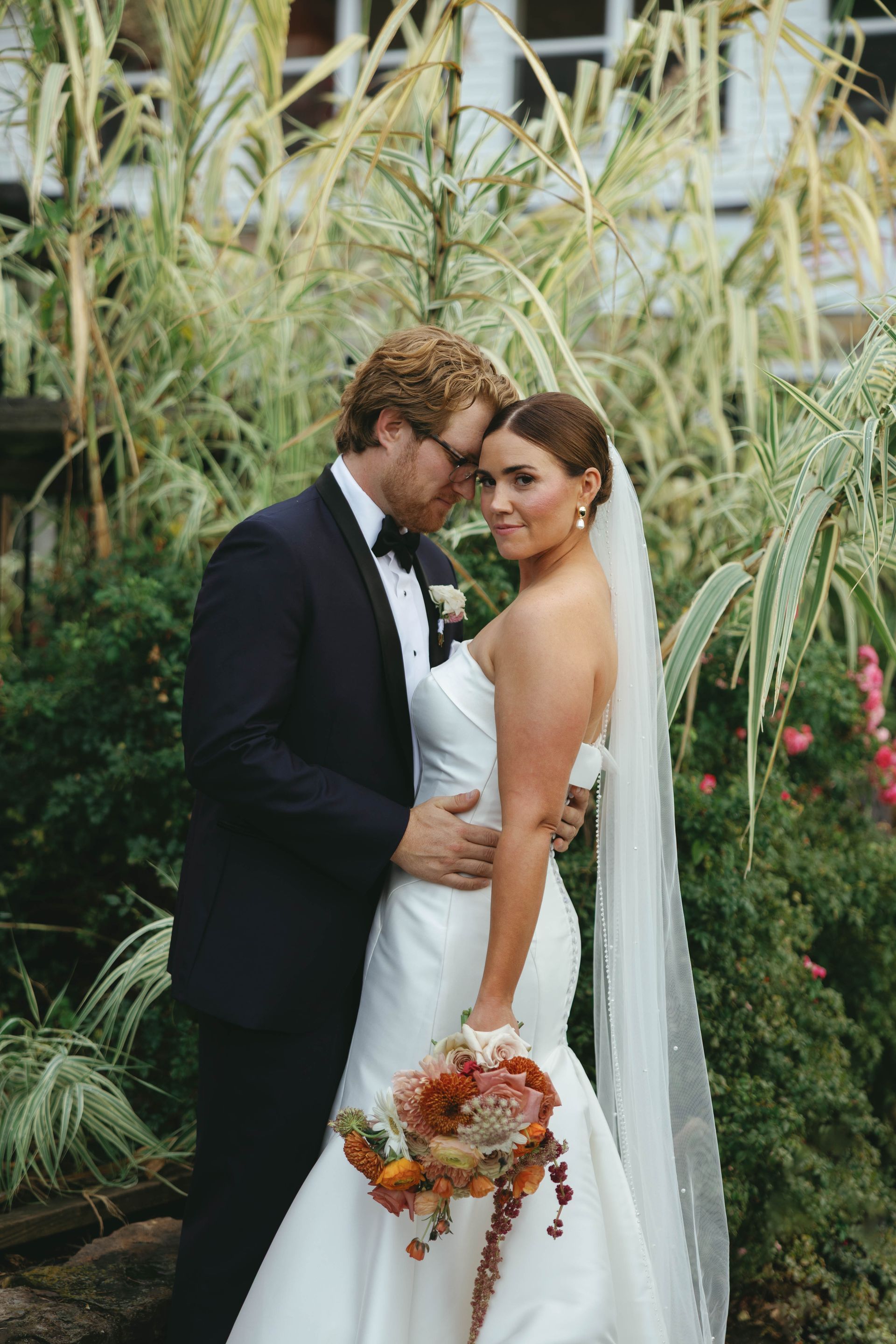 Bride and groom embracing, she in a white strapless dress, he in a suit with a bow tie, standing in front of greenery.