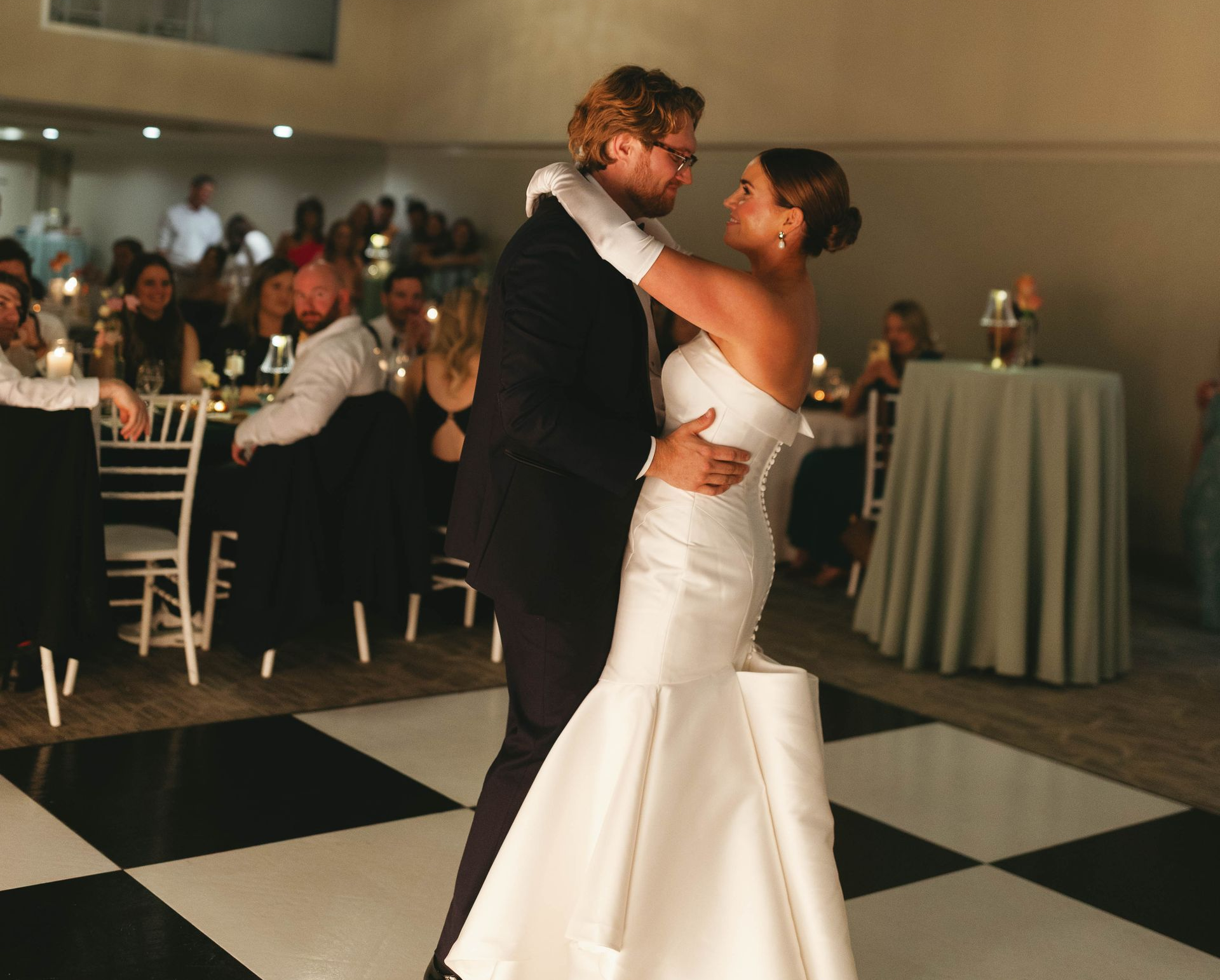 Bride and groom dance on a checkered floor at a reception. She wears a white gown, he wears a suit. Guests watch.