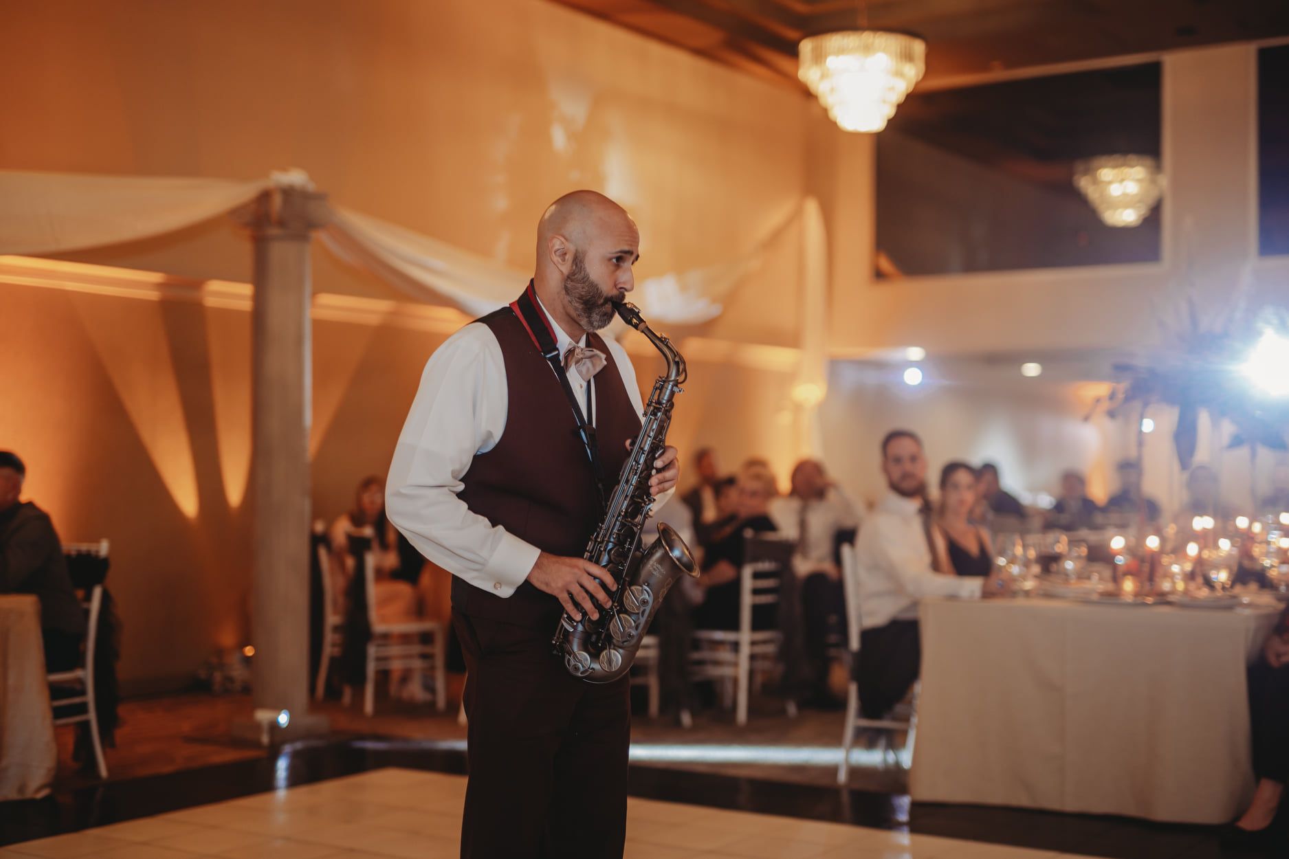 Saxophone player in a formal setting, performing.  Warm lighting, people seated at tables in the background.