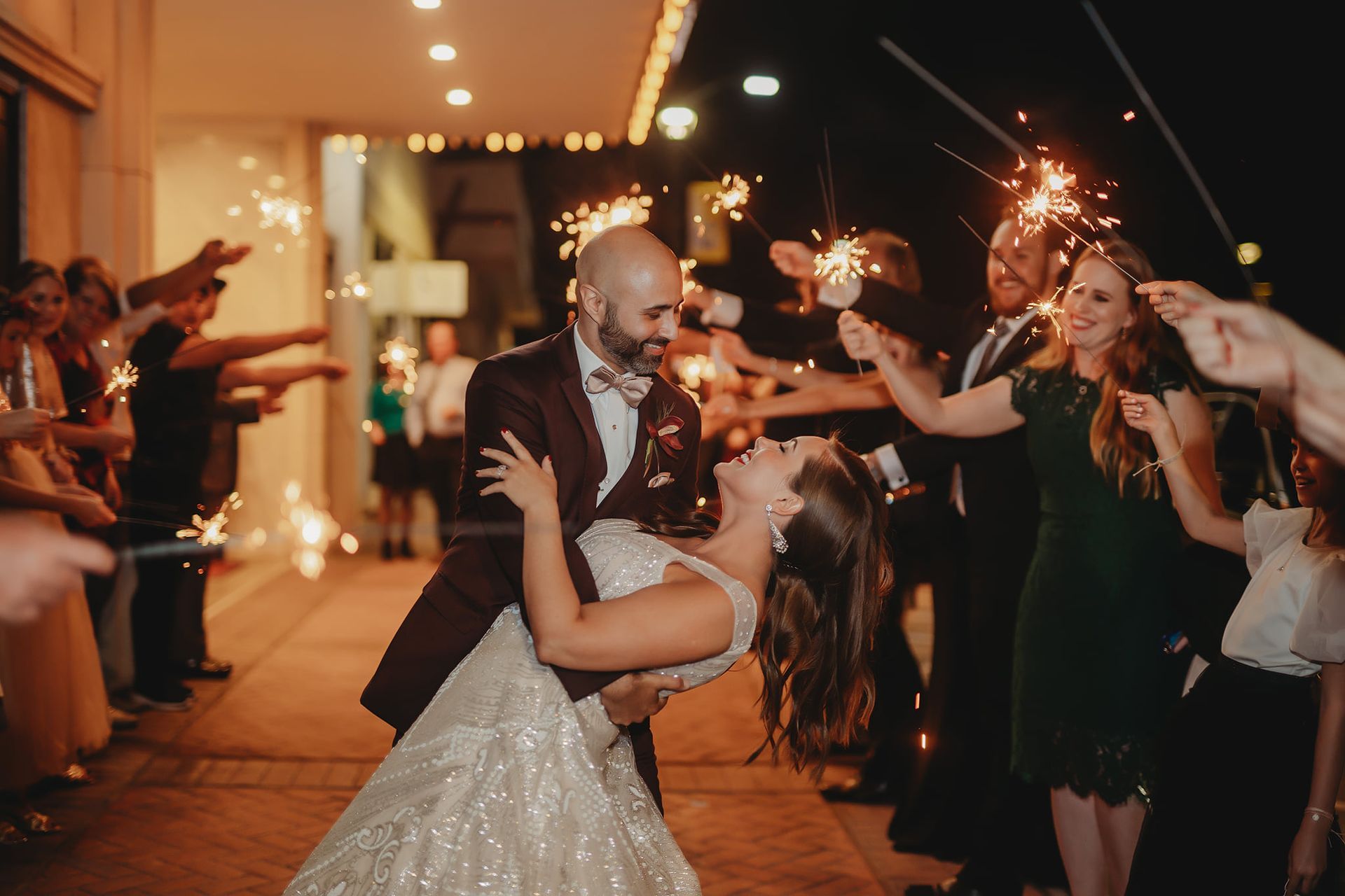 Bride and groom dip beneath sparklers as guests cheer at a night wedding celebration.