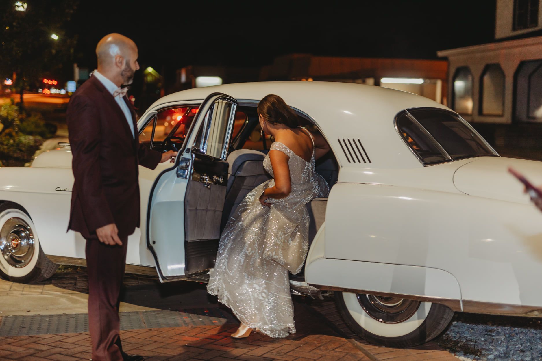 Man in suit assisting a woman in a silver dress exiting a white vintage car at night.