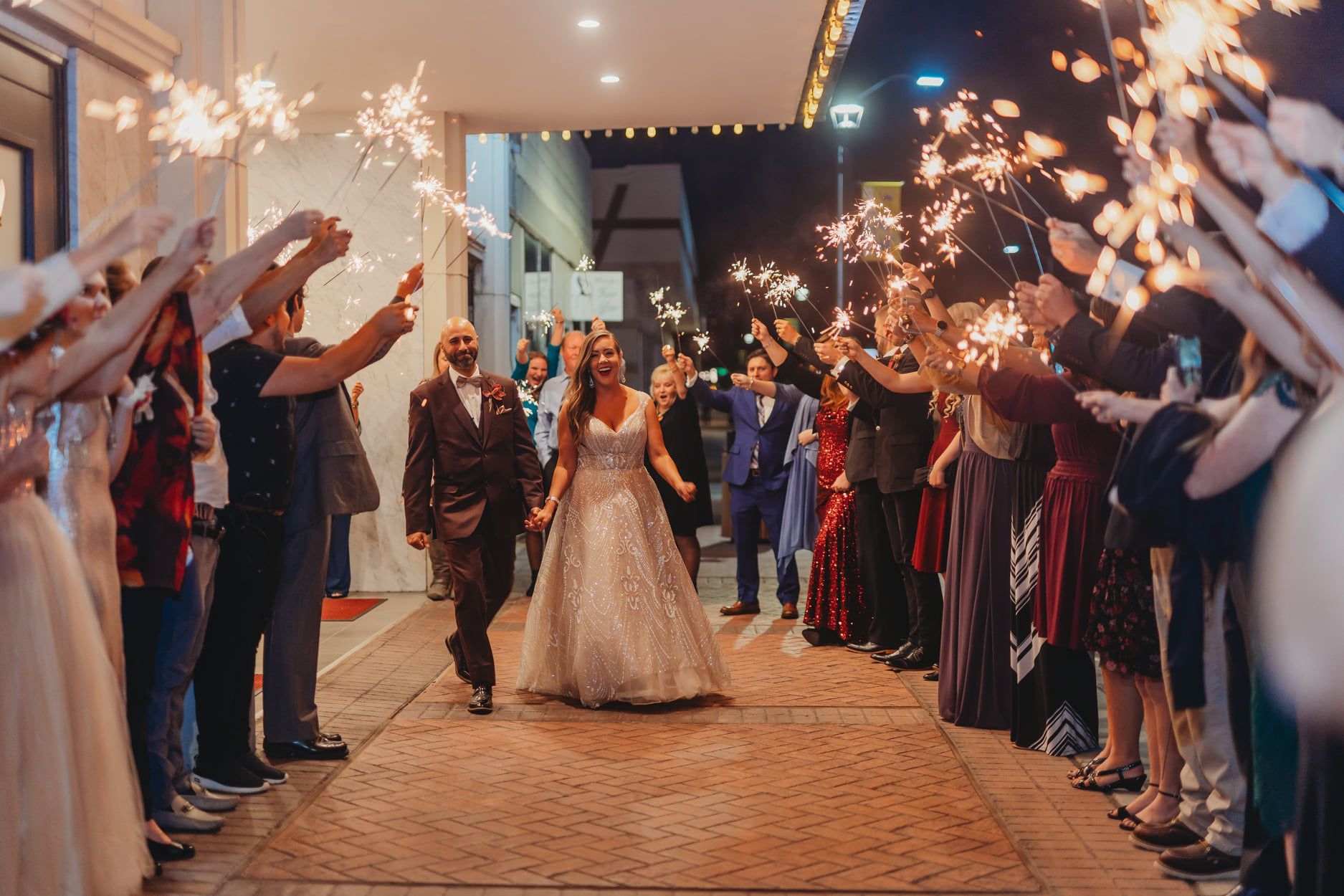 Newlyweds exit venue under a sparkler send-off; night, city backdrop. Guests cheer, holding sparklers.