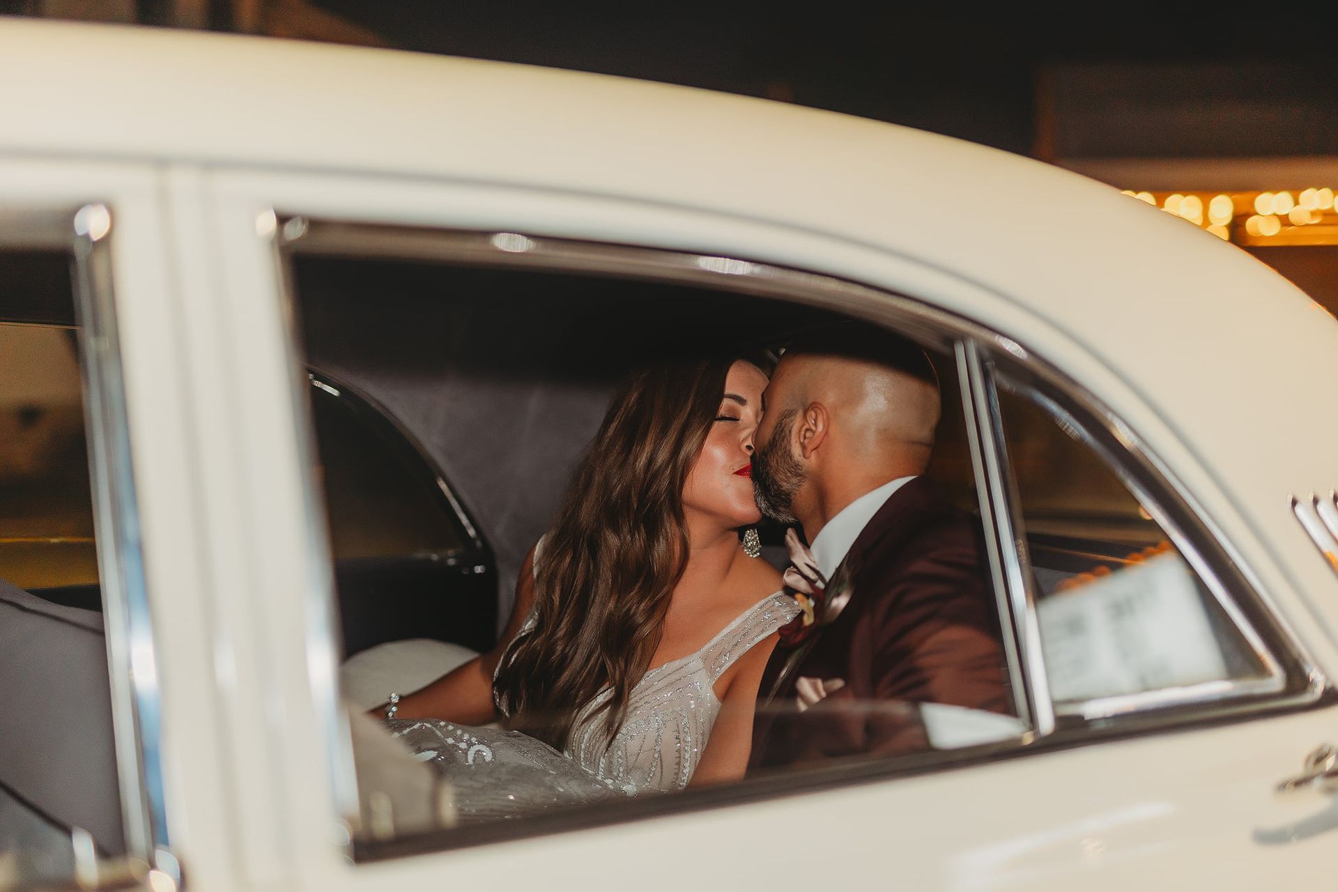 Couple kissing in a vintage car; bride wearing a sparkly gown, groom in a burgundy suit.