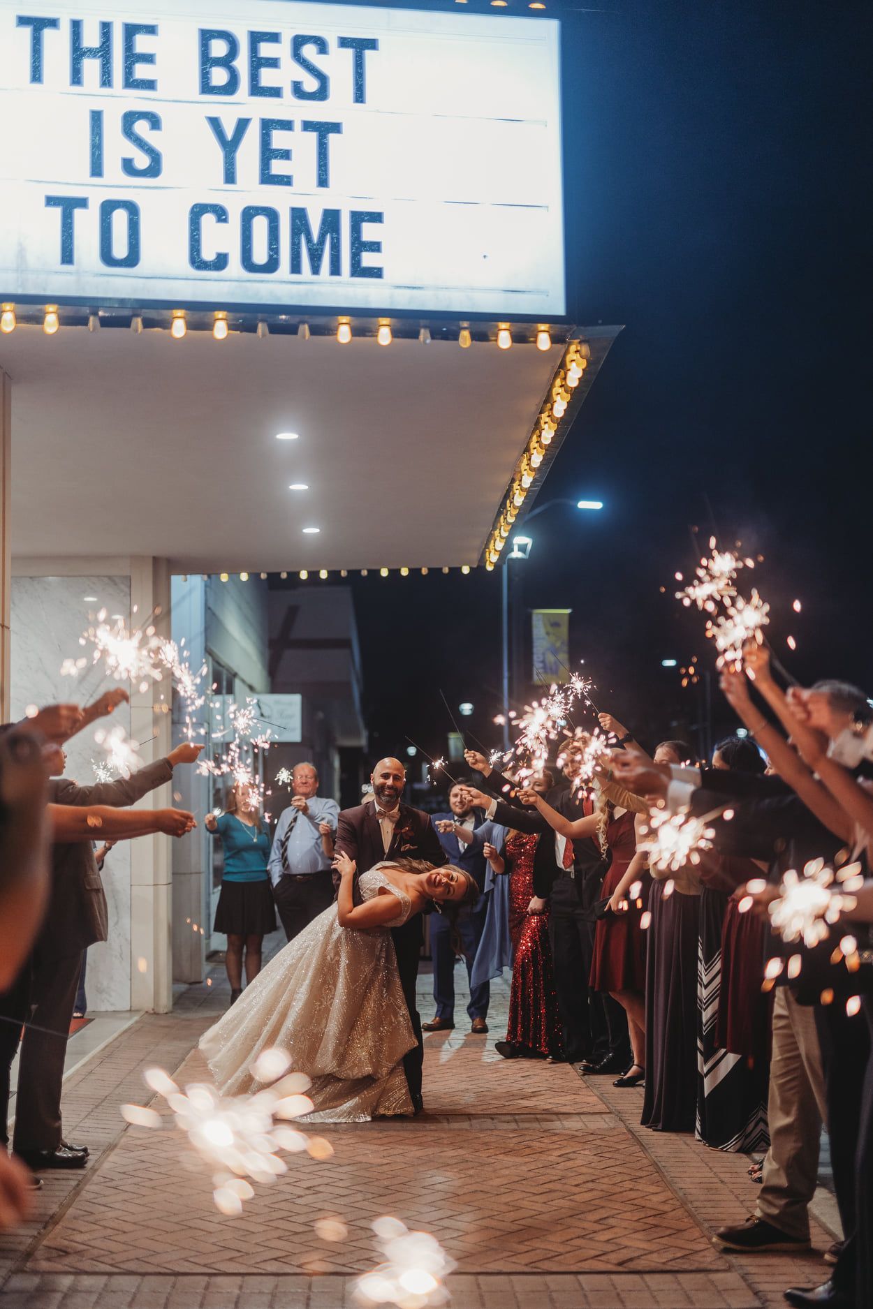 Couple dances under sparklers outside a theater at night, sign reads 
