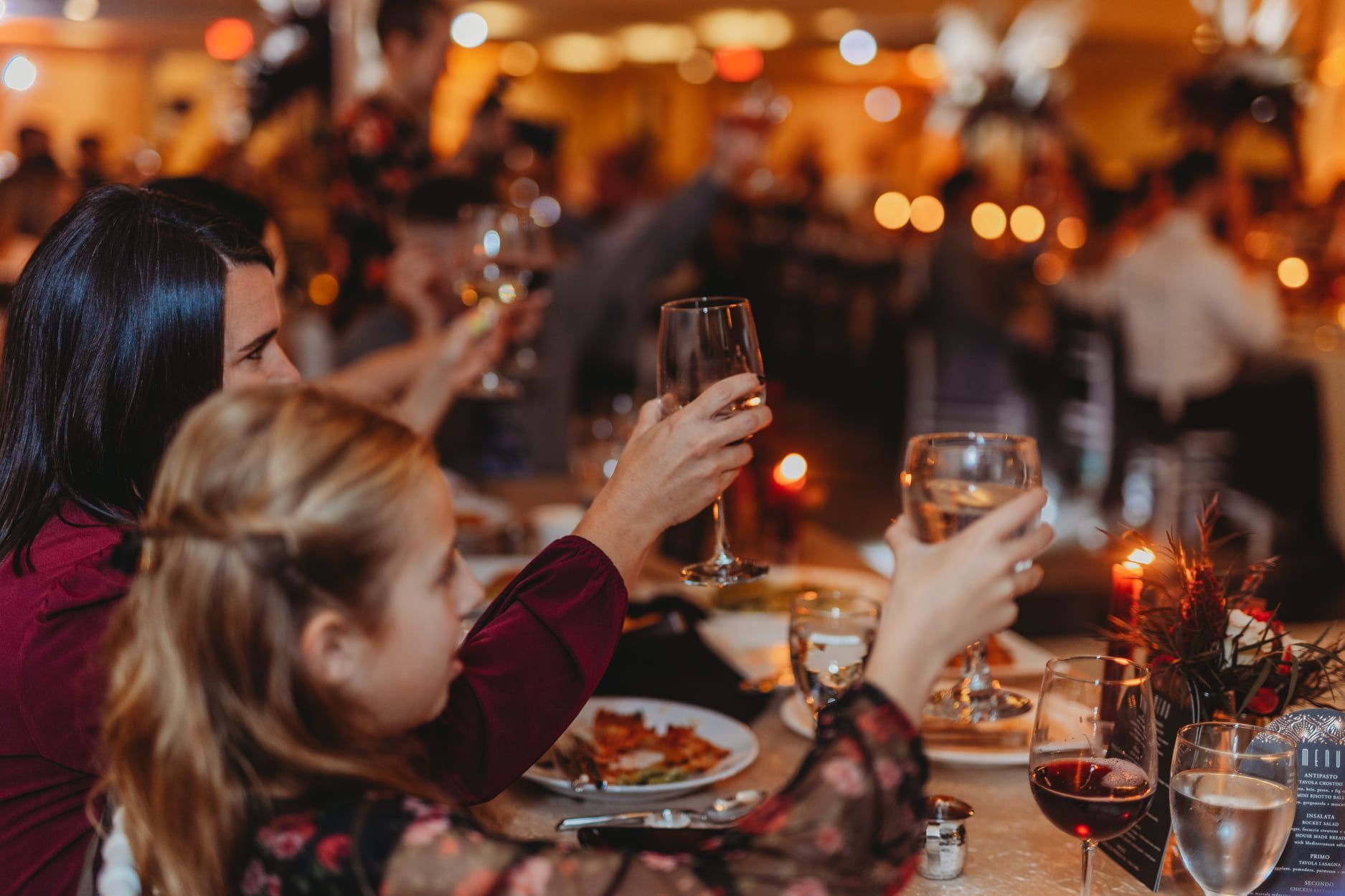 People at a table raise wine glasses. A young girl looks up, while others eat at a warmly lit gathering.