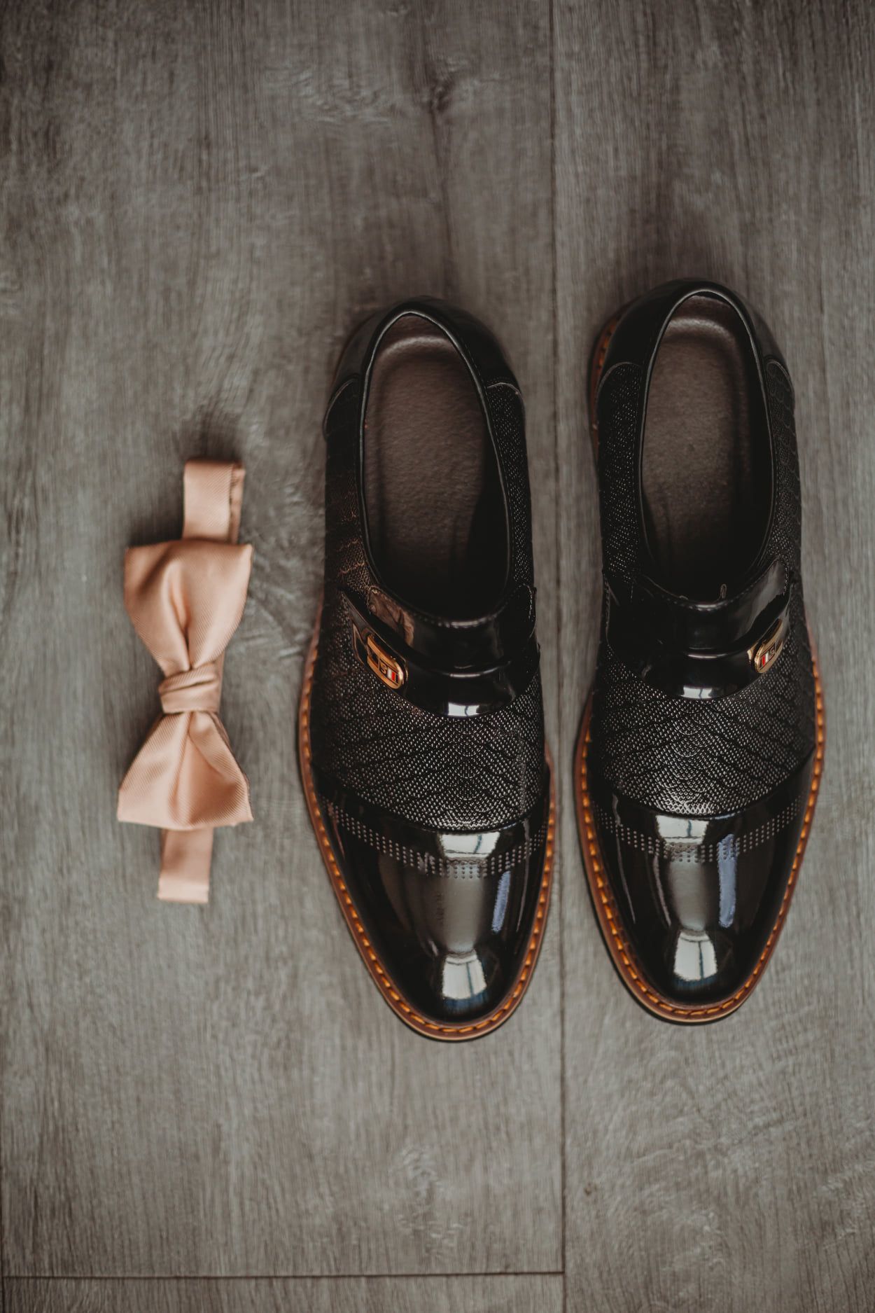 Black, shiny leather shoes and a light pink bow tie on a gray wood surface.
